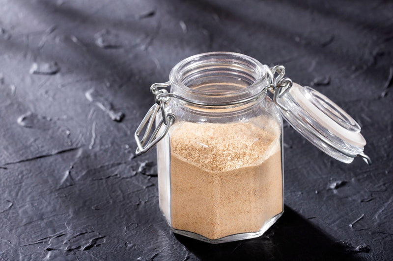 Glass jar filled with fine lion’s mane mushroom powder on dark textured surface with open clamp lid, highlighting texture and storage.