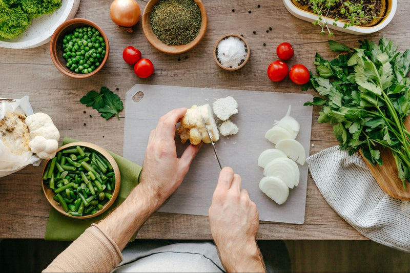 Overhead view of fresh Lion’s Mane mushroom being sliced on a cutting board surrounded by onions, peas, tomatoes, herbs and leafy greens.