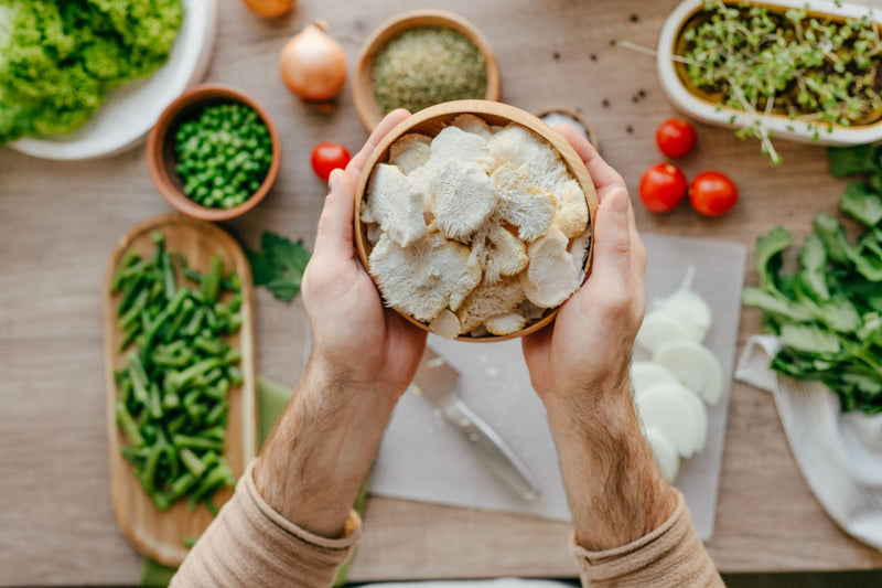 Overhead view of hands holding bowl of sliced Lion’s Mane mushrooms with fresh vegetables on kitchen table.